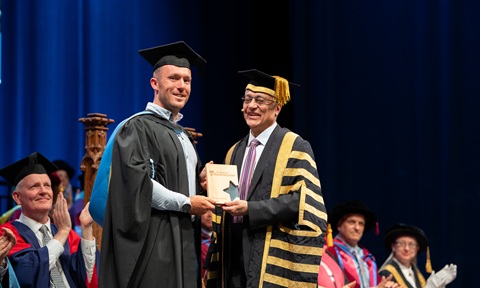 A graduate in academic dress receives an award from a university official wearing ceremonial robes.