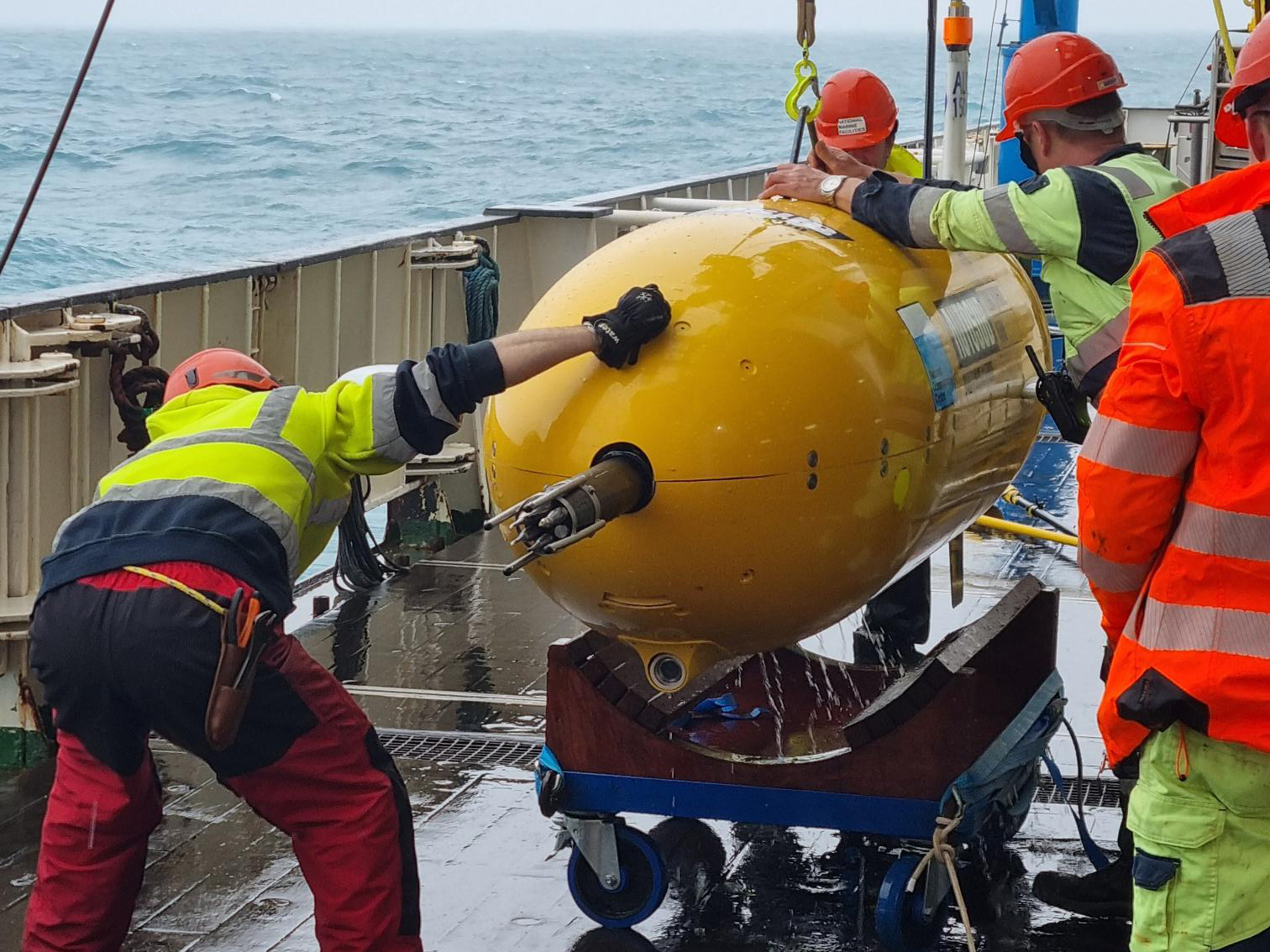 Yellow, tubular autonomous submarine being winched up from a wheeled platform, onboard a research vessel. The submarine is being inspected by four crew members wearing high visibility clothing and hard hats. Some have radios and toolbelts. The deck of the ship is wet and the ocean waves are visible in the background. 