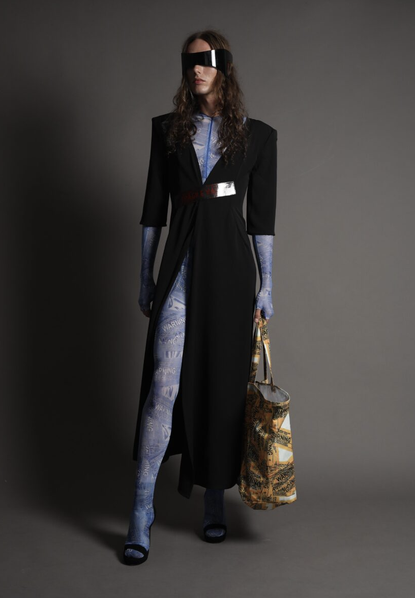 A person stands against a dark background wearing a futuristic outfit that includes a long black coat with metallic accents, a blue patterned bodysuit, and large visor-style sunglasses. They hold a large, intricately designed bag.