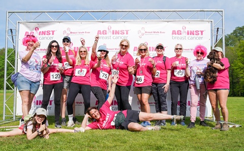 Group of 13 people outside on the grass standing in front of a large white banner stating Against Breast Cancer. Most of the group are wearing pink T-shirts