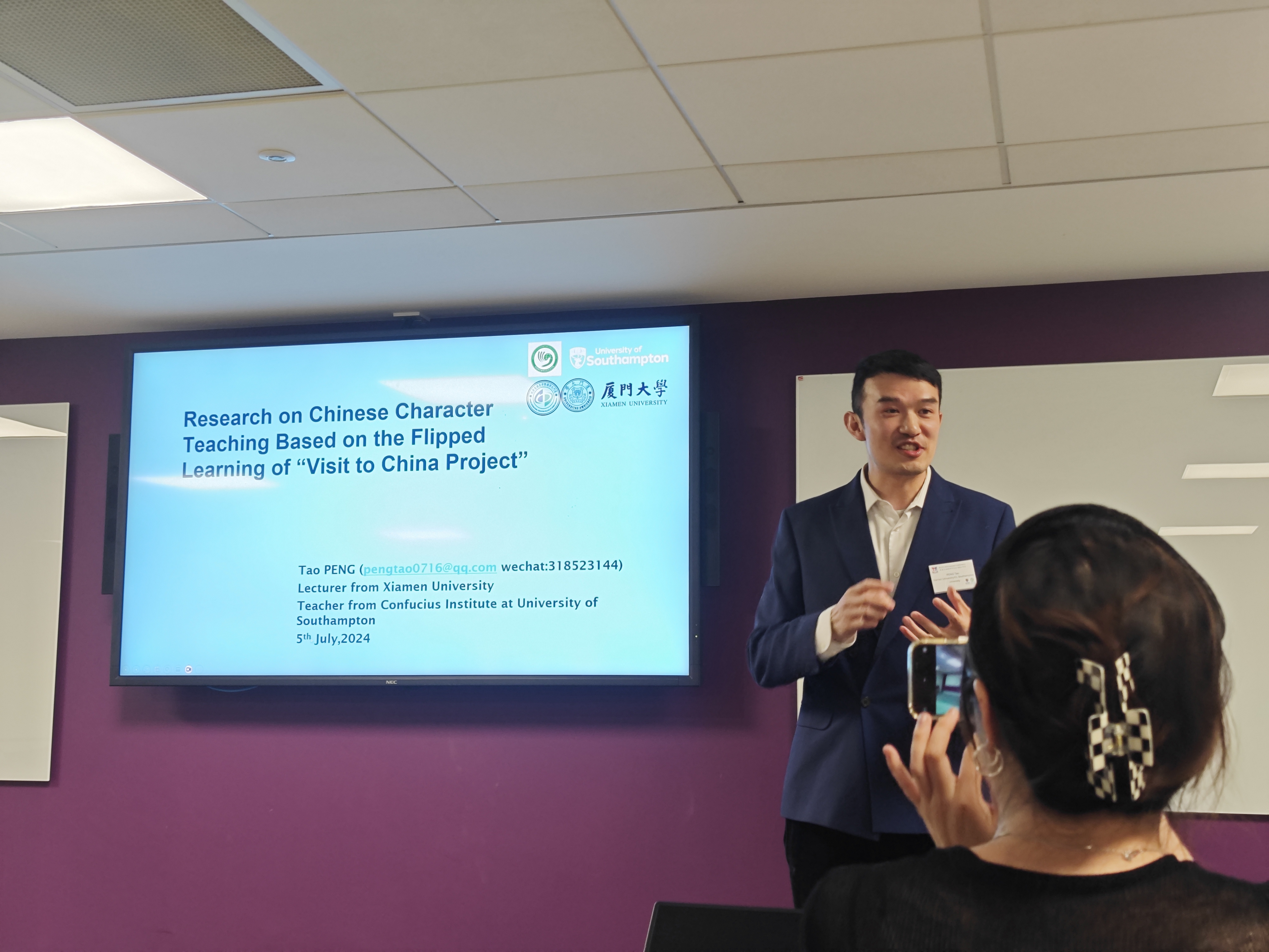 British Chinese Language Teaching Society lecturer with presentation scren behind. A member of the audience is taking a photography