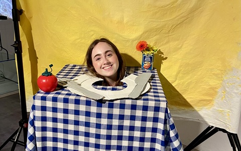 A young woman is inside a sculpture of a dining table with a plate on it, and her head is sticking up through the plate. 