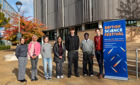 Seven people standing in a line facing the camera, with a blue banner on the right headlined 'British Science Festival'. They are stood on a paved area in front of a large wood panelled buiding 
