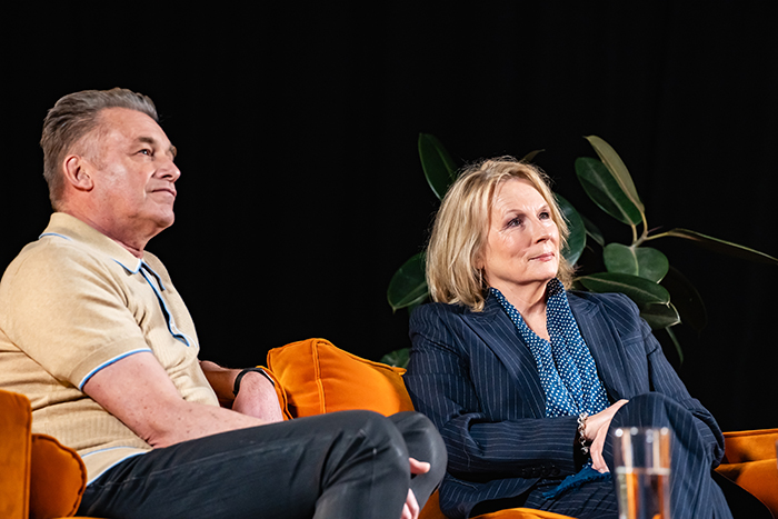 Two people sit on orange velvet chairs on a stage against a black backdrop, appearing to listen during a panel discussion or interview. A man, Chris Packham, in a beige polo shirt sits on the left, while a woman, Jennifer Saunders, in a navy pinstripe suit and blue patterned blouse sits on the right. A leafy green plant and a glass of water are visible in the background and foreground.
