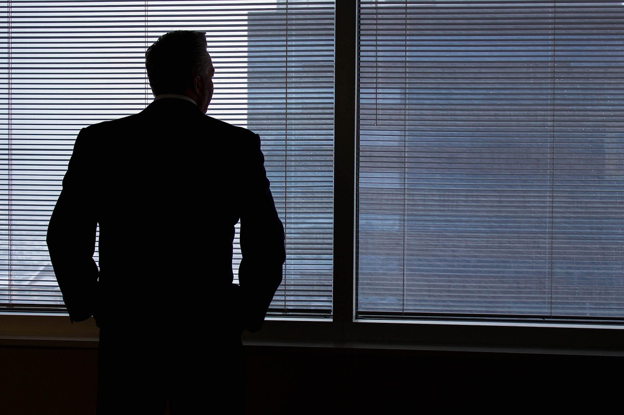 A silhouette of a man in a suit standing near a window with horizontal blinds, gazing outside. The scene suggests a contemplative or reflective moment, with a muted urban or office setting visible through the blinds.