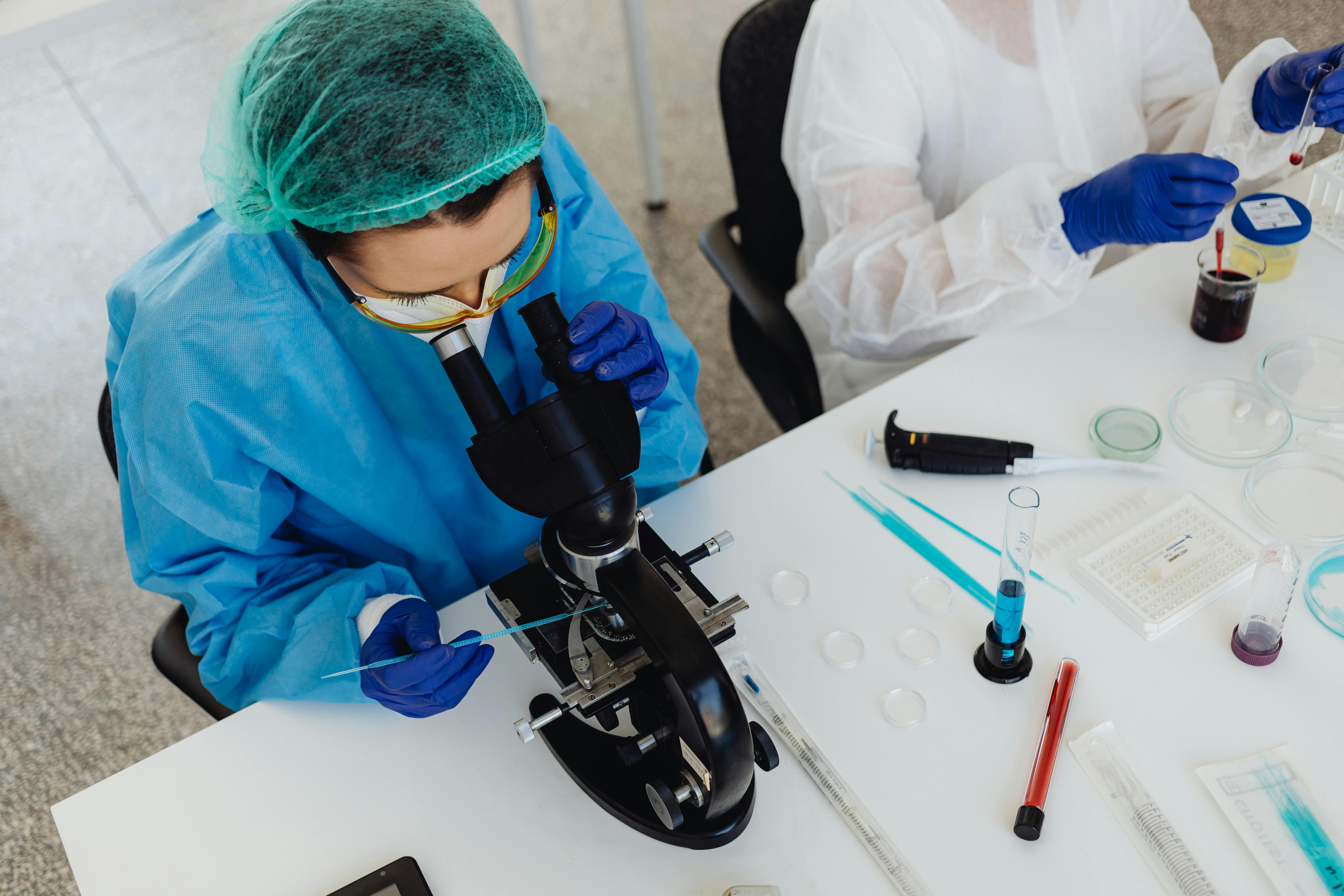 Scientist in lab wearing blue gown, headwear and yellow glasses looking into microscope