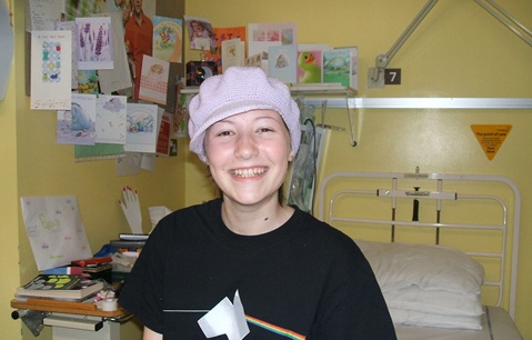 Teenage girl wearing a knitted pale pink beret and a black T-shirt, smiling. She is stood in a hospital ward with a hospital bed and a shelf full of greetings cards behind her