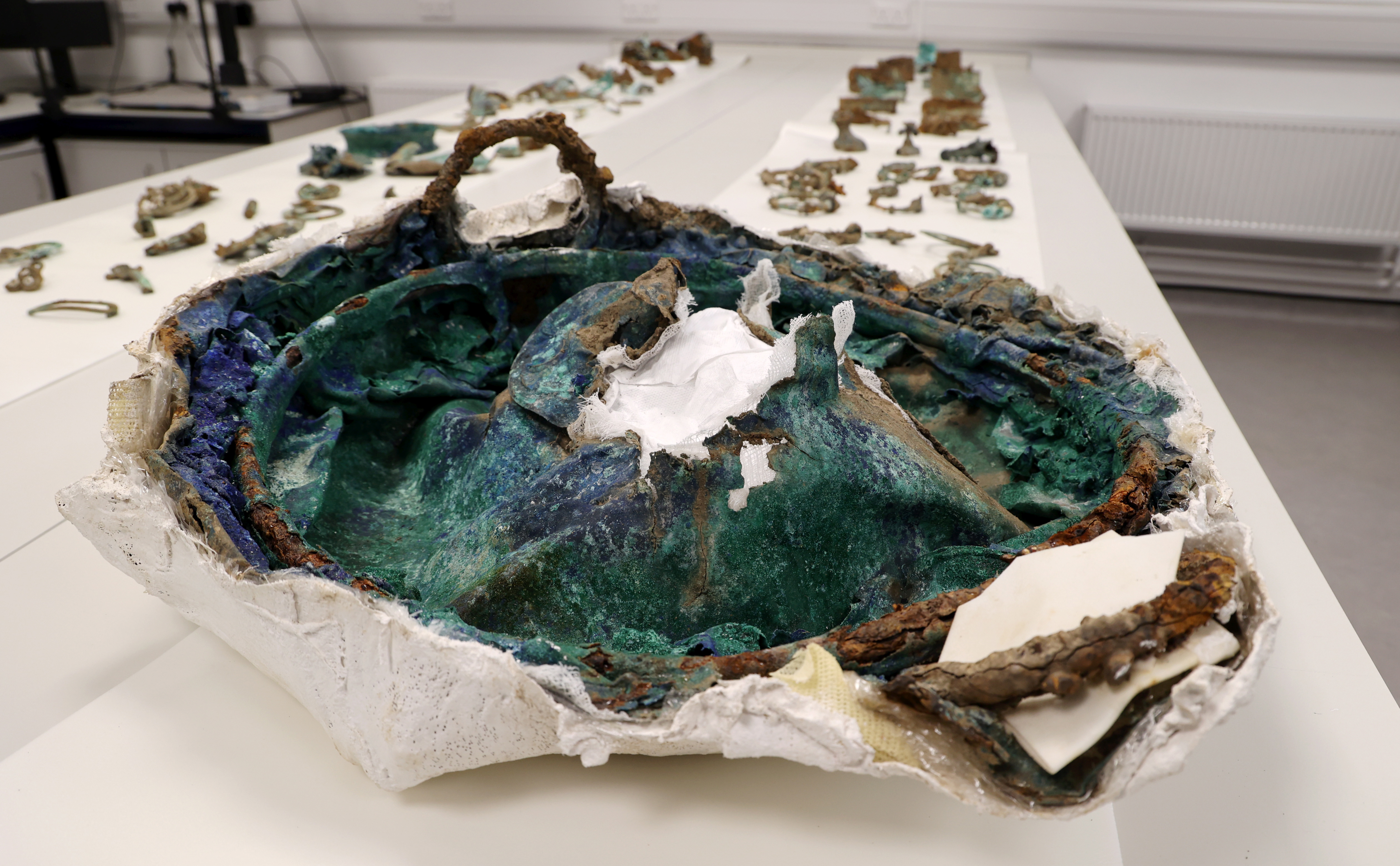 Ancient cauldron, green in colour and only partially intact, laying on a table in front of other archaeological artefacts.