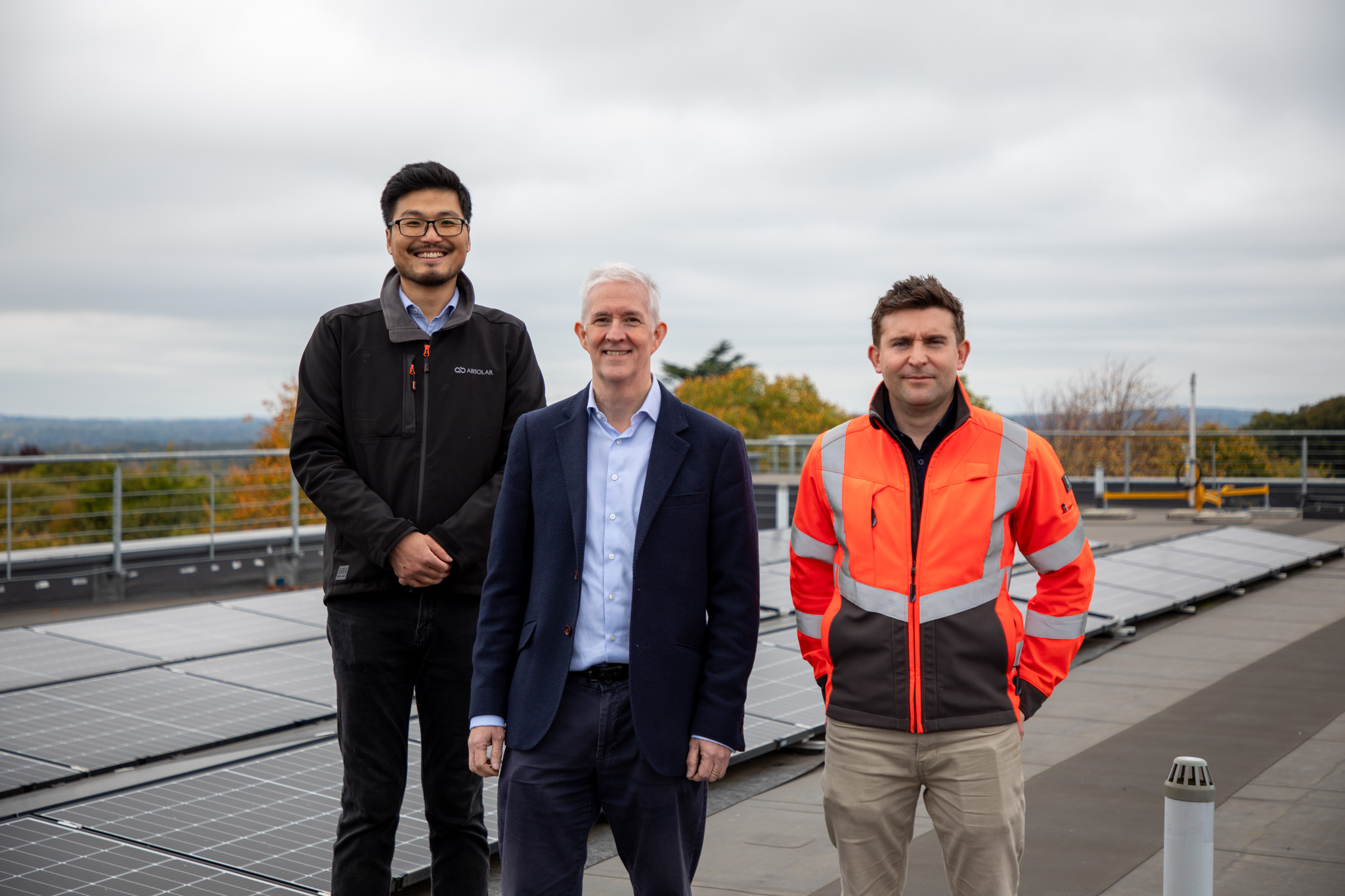 Three people are standing on the roof of a building. Solar panels and trees can be seen behind them.