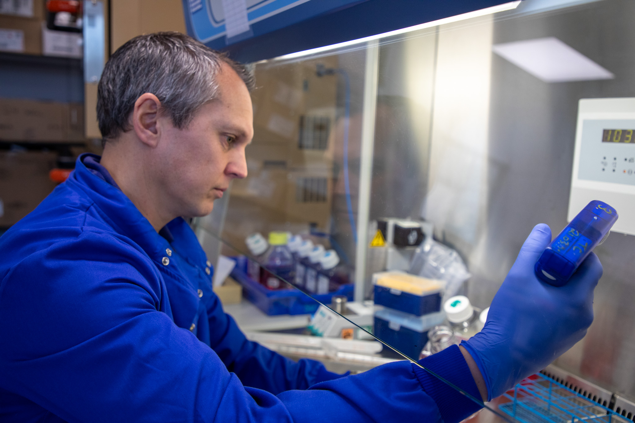 A scientist in a blue lab coat and blue gloves works inside a biosafety cabinet. He is holding a handheld device while looking intently at laboratory equipment and containers inside the cabinet. Bottles, tubes, and lab supplies are visible around him.