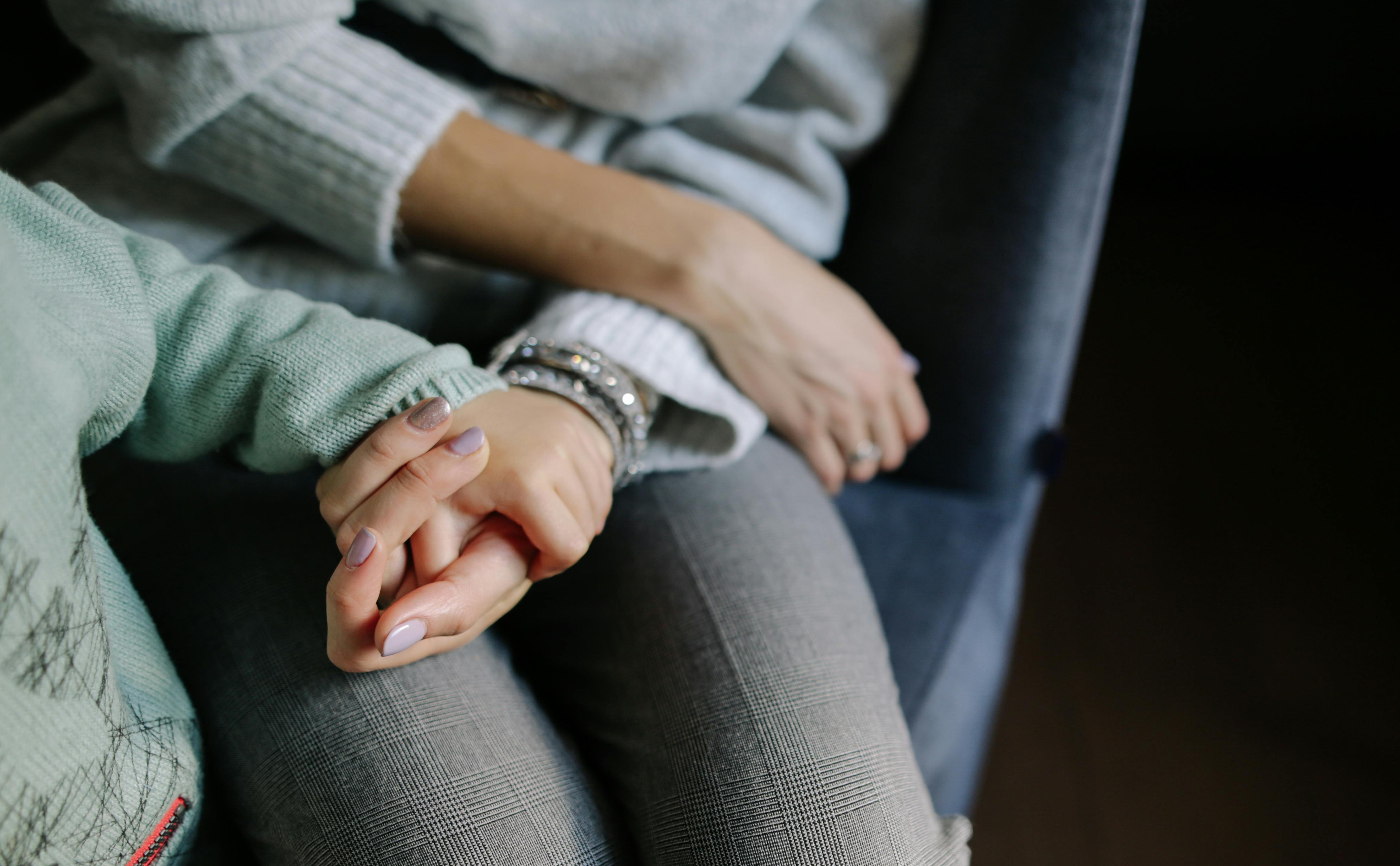 A woman's hand cupped upwards holding a toddler's hand with both rested on the woman's leg.