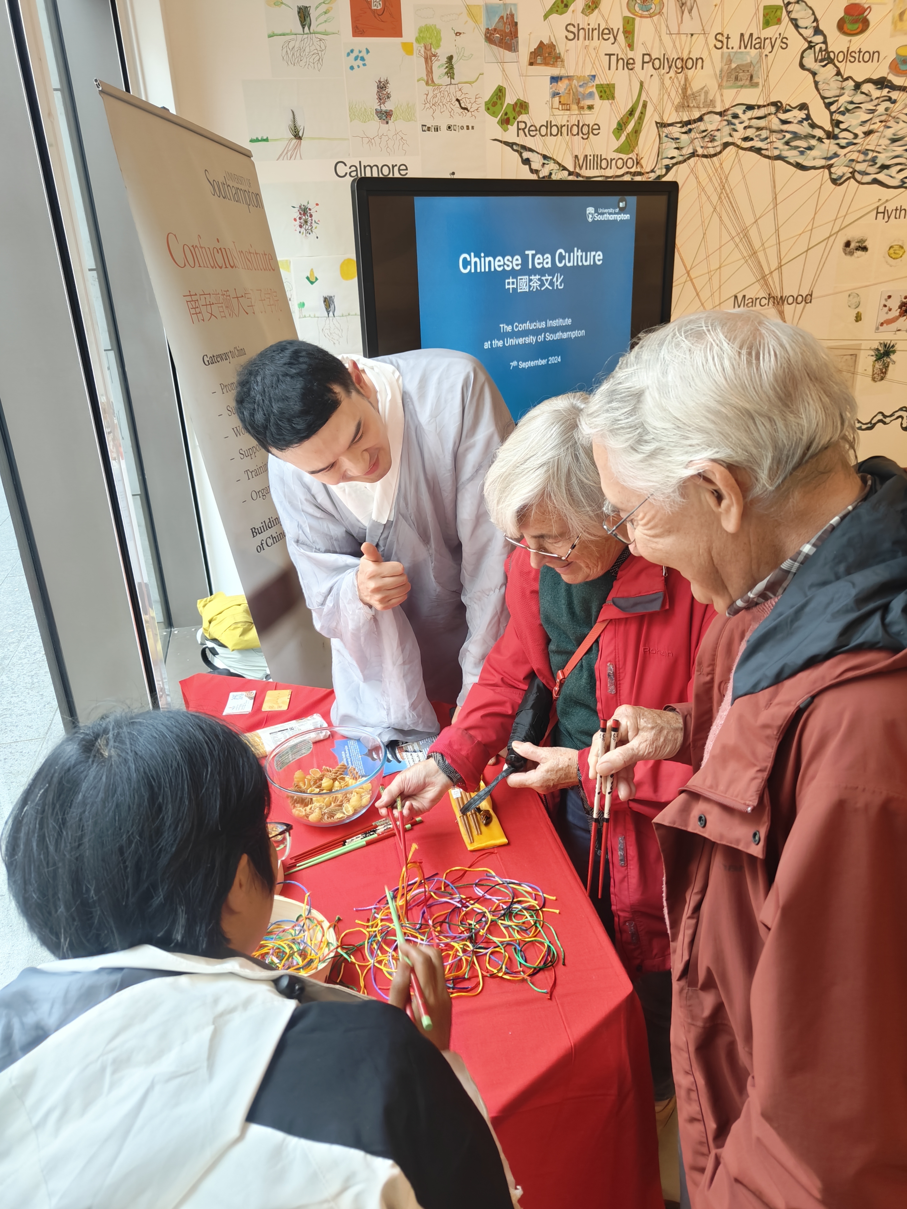 A group of people gathers around a table covered with colorful strings and chopsticks, engaged in an activity at a Chinese tea culture event. One individual in traditional Chinese clothing is instructing the group, with a screen in the background displaying "Chinese Tea Culture" hosted by the Confucius Institute.