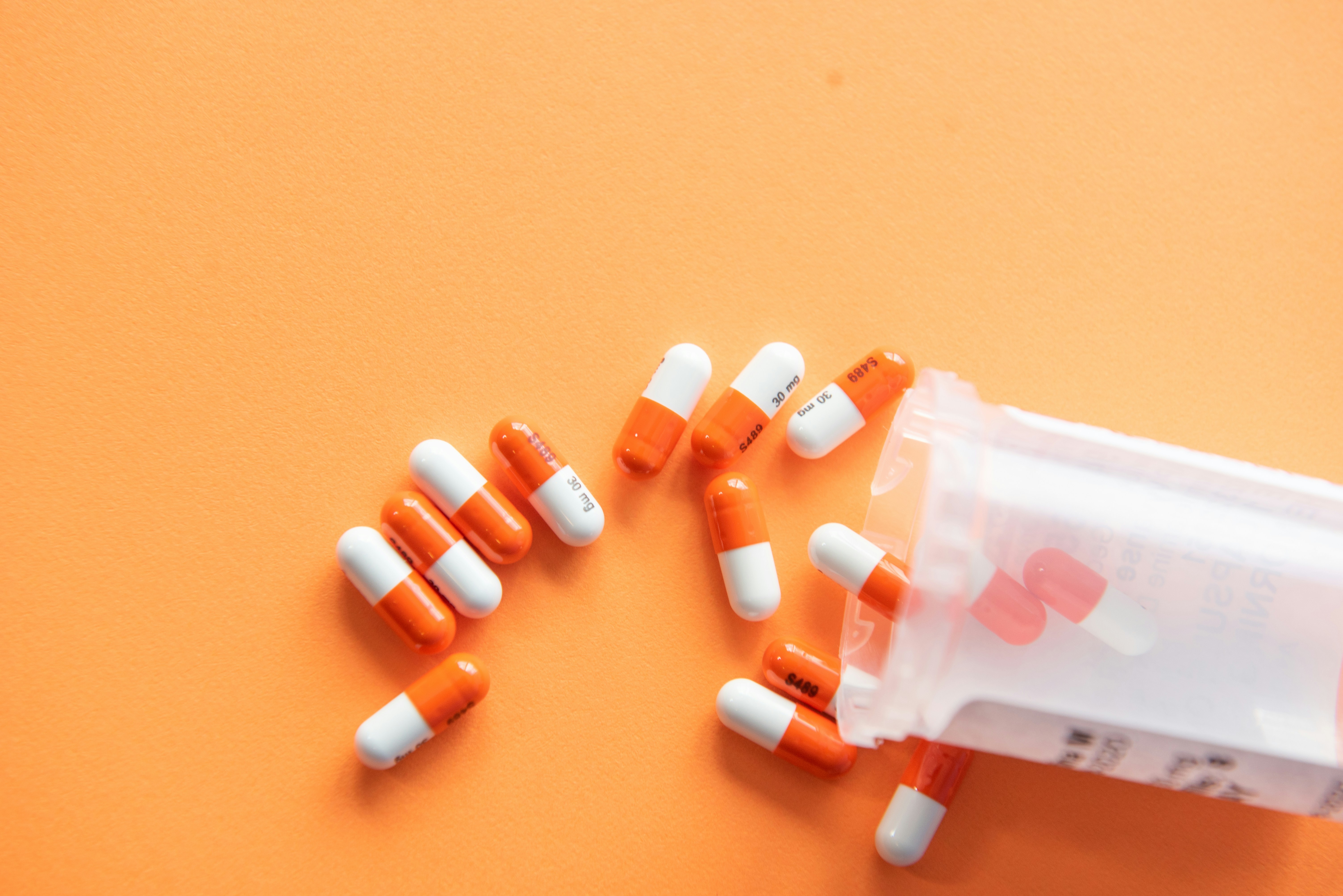 An open prescription bottle on its side with orange-and-white capsules spilling out onto a solid orange surface.