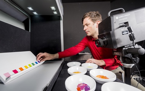 A man in a red long sleeved top looking at small tiles with different colours of paint on them. He is sat insude in a darkened room. There are small bowls of paint on the desk in the foreground