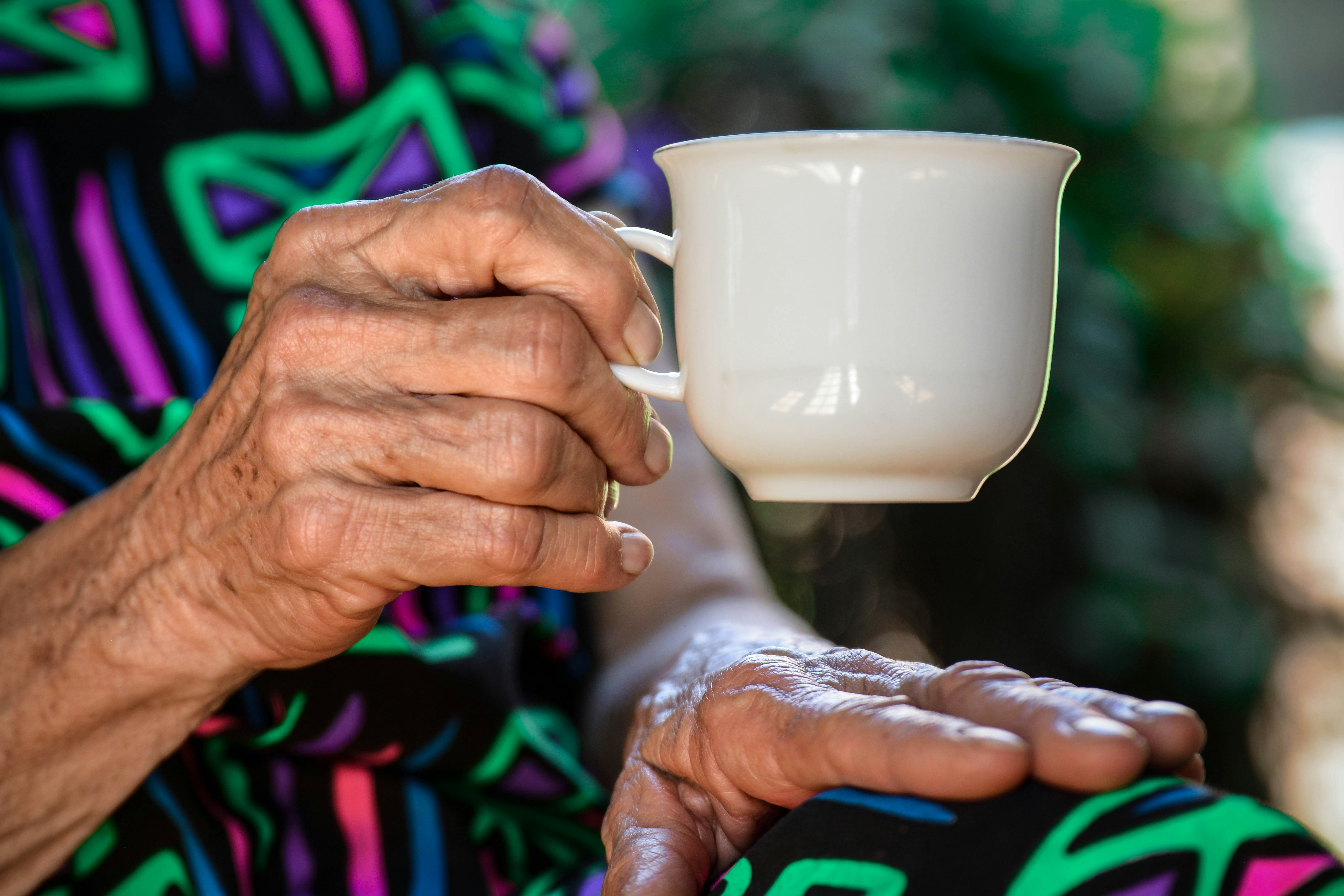 Close-up of an elderly person's wrinkled hands, one holding a white teacup and the other resting on their lap. The person is wearing colorful patterned clothing with green, purple, blue, and pink designs.