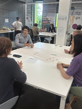 People collaborate around tables in a bright classroom or workshop space with papers, posters, and greenery visible through the windows.