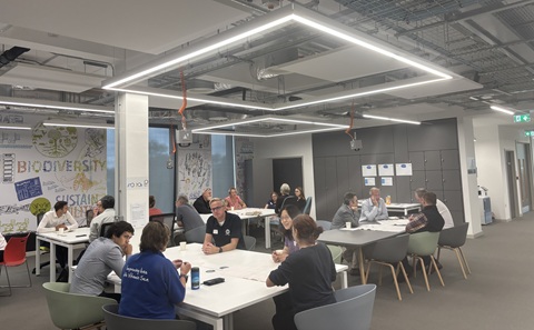 People collaborate around tables in a modern, brightly lit meeting room with sustainability-themed wall artwork.