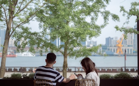Rear view of a couple sat on a part bench. One of them is looking at their phone 