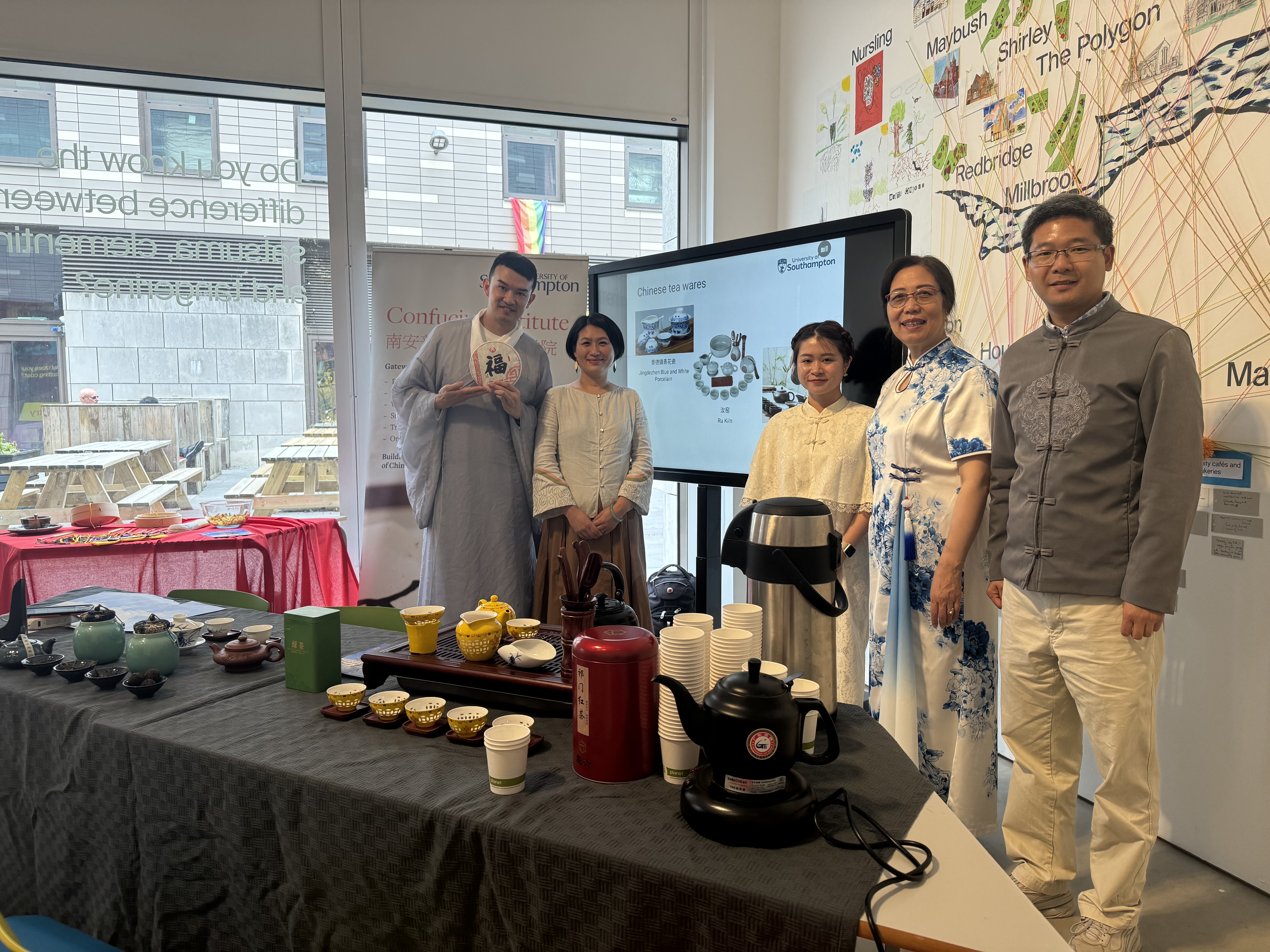 A group of five people, dressed in traditional Chinese clothing, stand together next to a display of tea sets at an indoor cultural event. Behind them, there is a screen with information about Chinese tea wares and a map on the wall, while tables are arranged with various tea pots, cups, and thermoses.