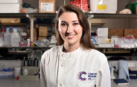 Woman with long dark hair wearing a white lab coat with the Cancer Research UK logo stitched onto it