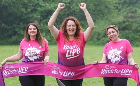 Three women in running gear and Race For Life T-shirts with a Race For Life banner