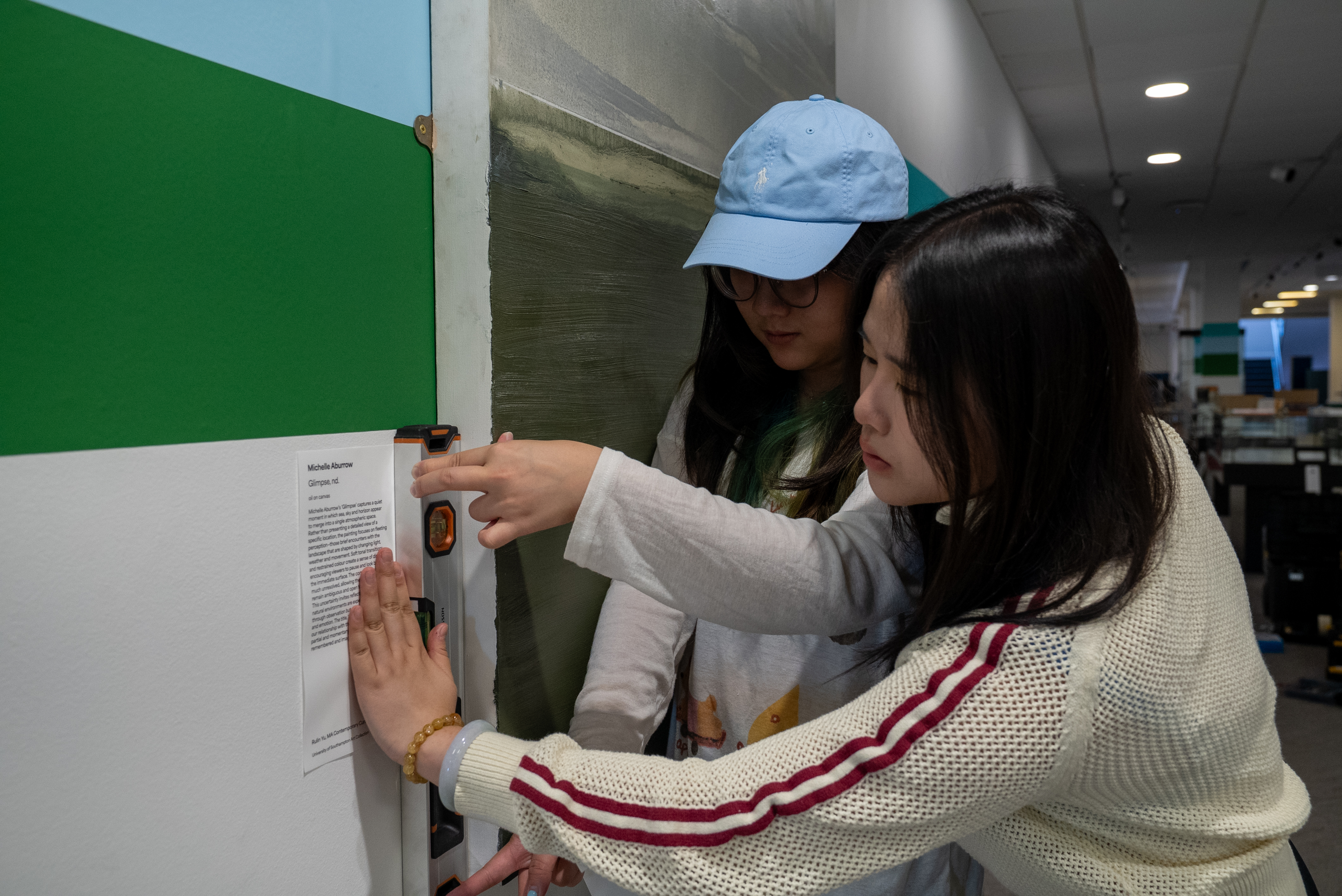 Two young women mounting an art description onto a wall next to a painting.