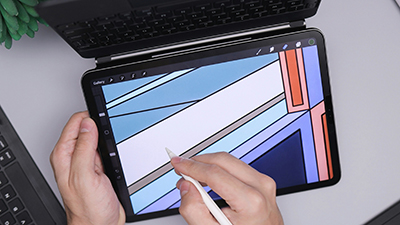 A person uses a stylus to draw a colourful geometric illustration on a tablet at a desk. A laptop is visible behind the tablet, with part of a plant in the corner.