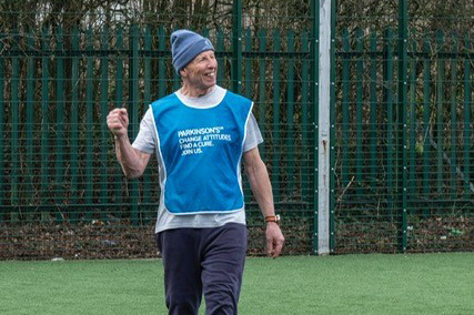 An older man is walking on an artificial turf football pitch, smiling and raising his fist in a celebratory gesture. He is wearing a blue sports bib over a grey t-shirt and a blue beanie hat. The bib has white text that reads: "PARKINSON’S CHANGE ATTITUDES. FIND A CURE. JOIN US." 