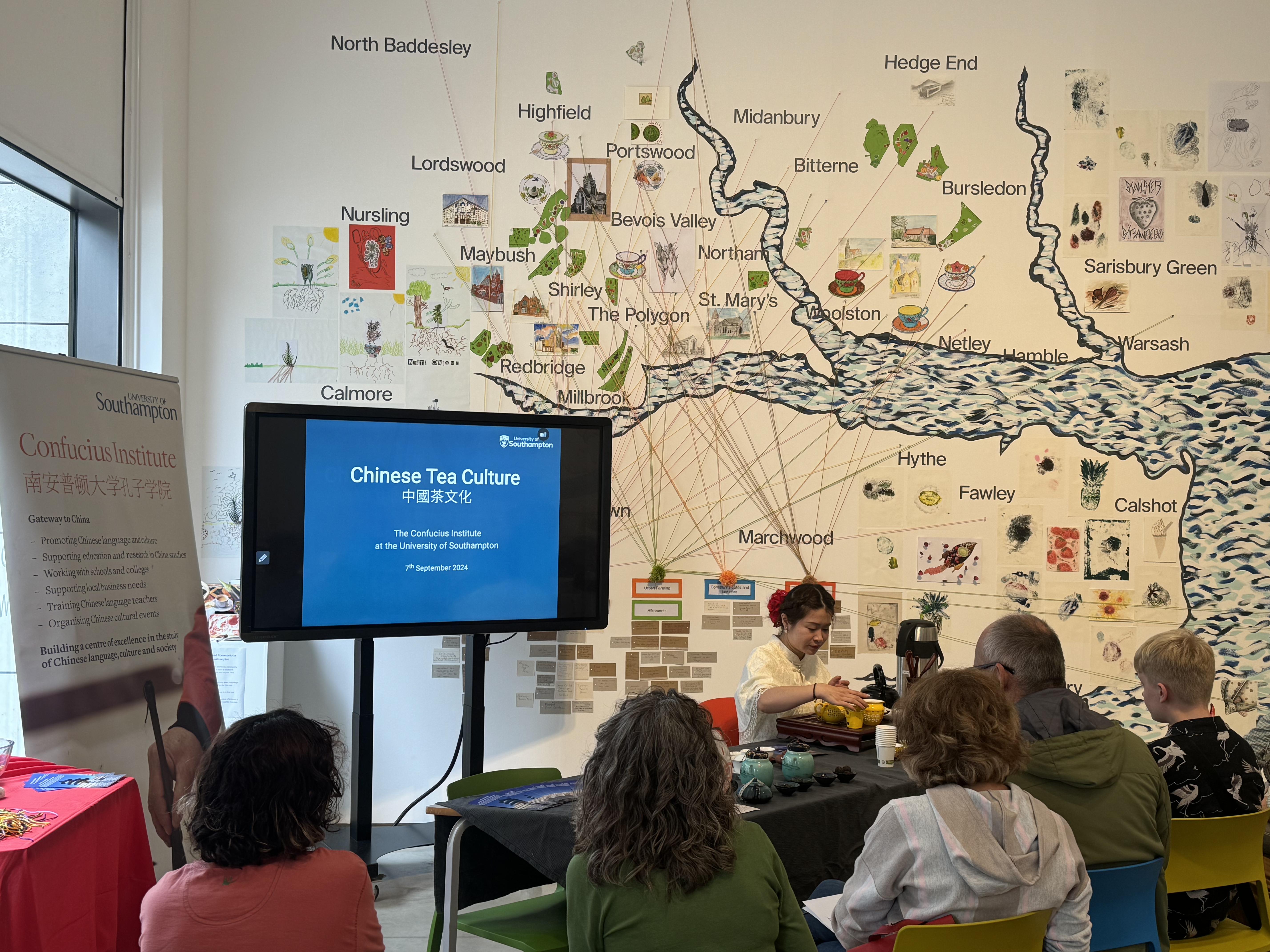 An audience sits in front of a presenter demonstrating Chinese tea-making techniques at an event, with a screen displaying "Chinese Tea Culture" hosted by the Confucius Institute. The background features a colorful map and drawings, while a table is set with tea sets and related items.