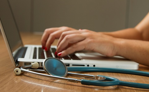 Close up photo of a person's hands at a computer keyboard, with a stethoscope on the desk in the foreground 
