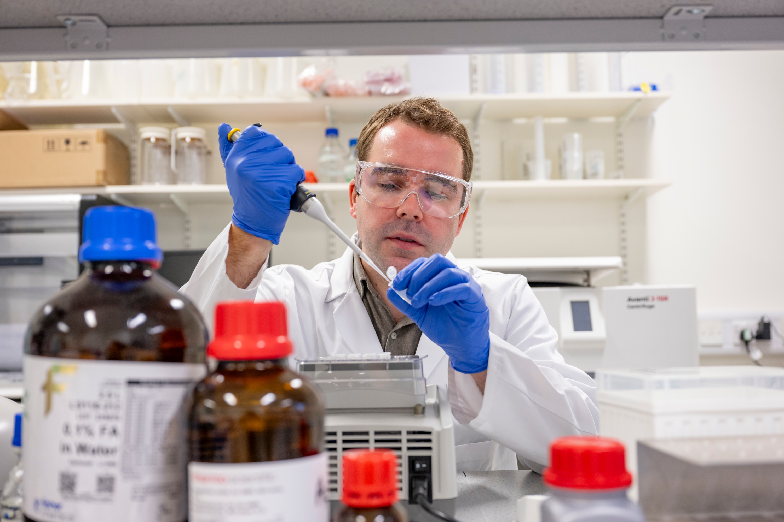 Person in a lab coat uses a pipette in a laboratory with shelves and bottles in the background.