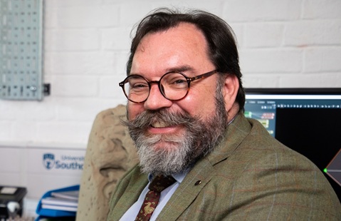 Head and shoulders photo of a man wearing a blazer, shirt and tie, with glasses and a beard, smiling. He is sat indoors, there is a computer monitor and a white brick wall in the background. 