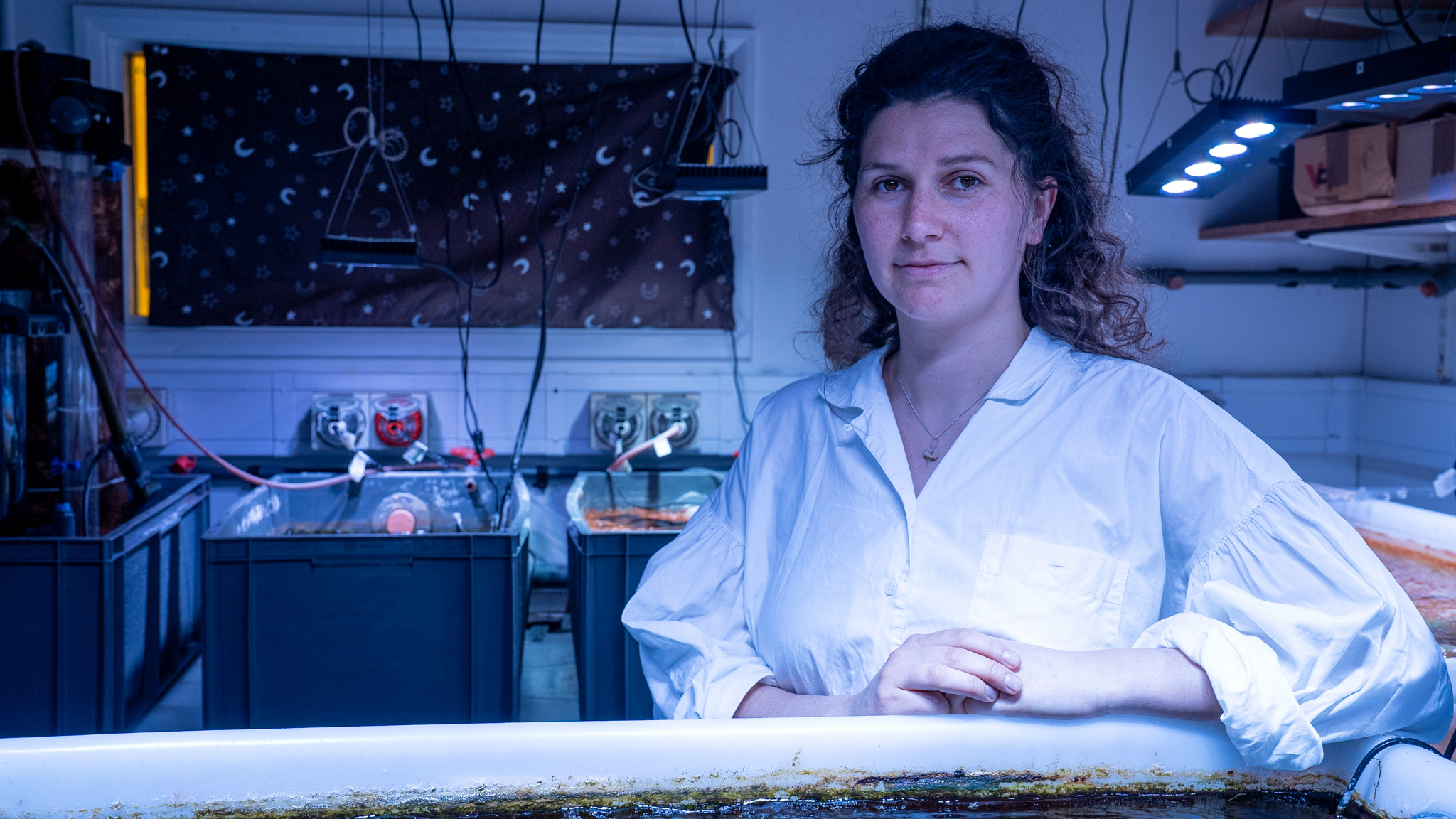 A woman wearing a white lab coat in the blue light of a laboratory aquarium.