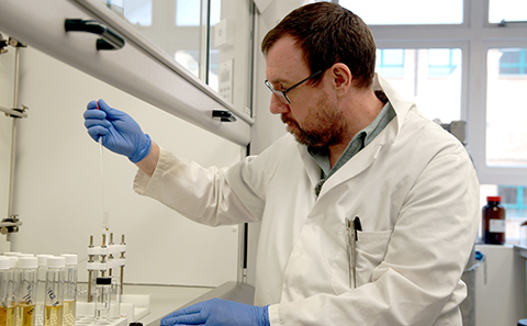 A side-profile of a man with glasses and a beard, wearing a white lab coat and blue gloves, using a pipette to carefully drop liquid into a glass vial within a laboratory fume hood.