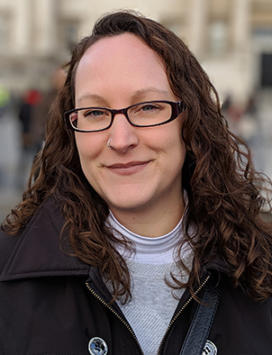 Head and shoulders image of a woman with long brown hair, wearing glasses and a dark jacket.