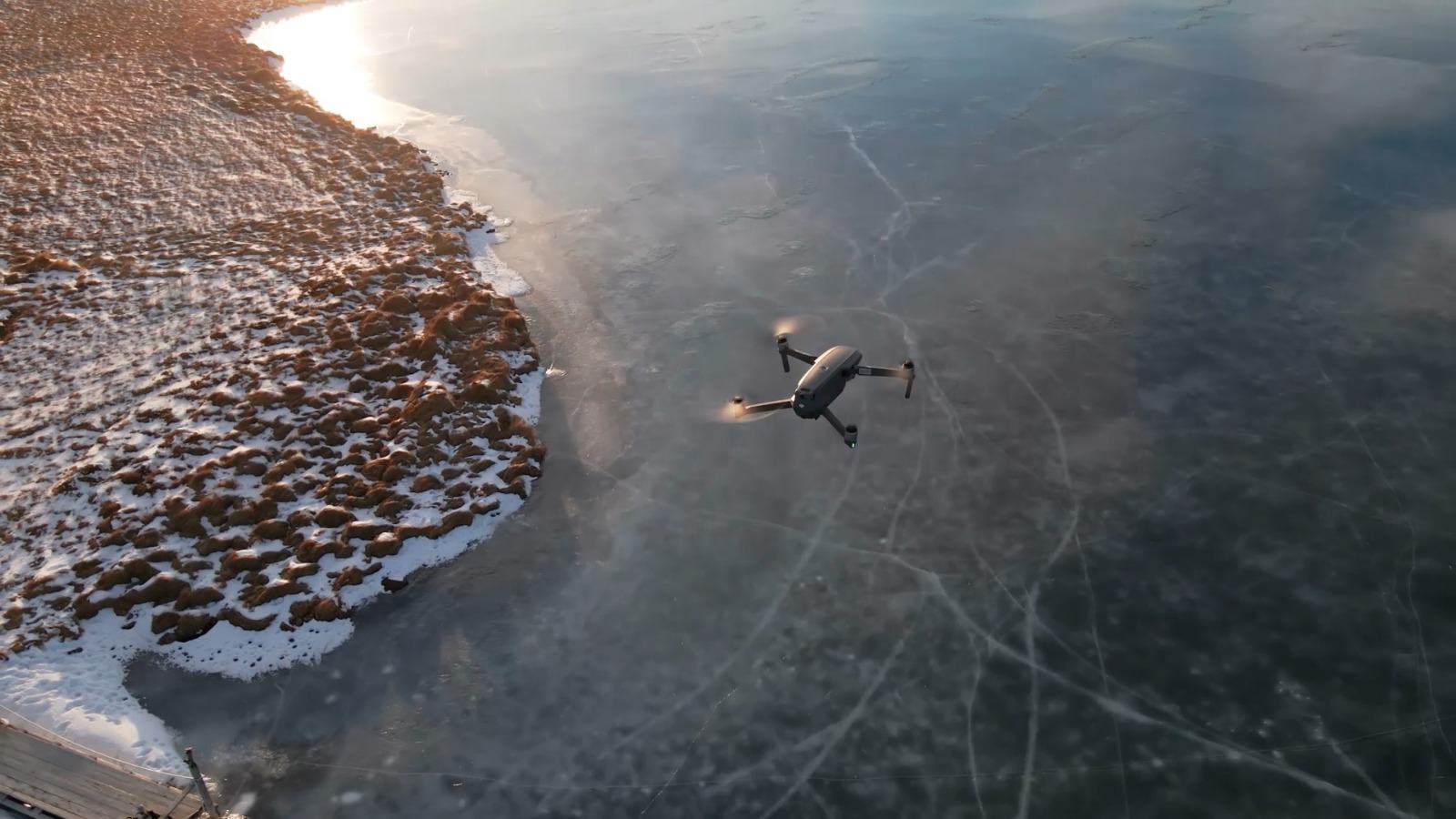 A drone flying over a frozen lake near a snow-dusted shoreline, with the low sun reflecting on the icy surface and patterns of cracks visible across the ice.