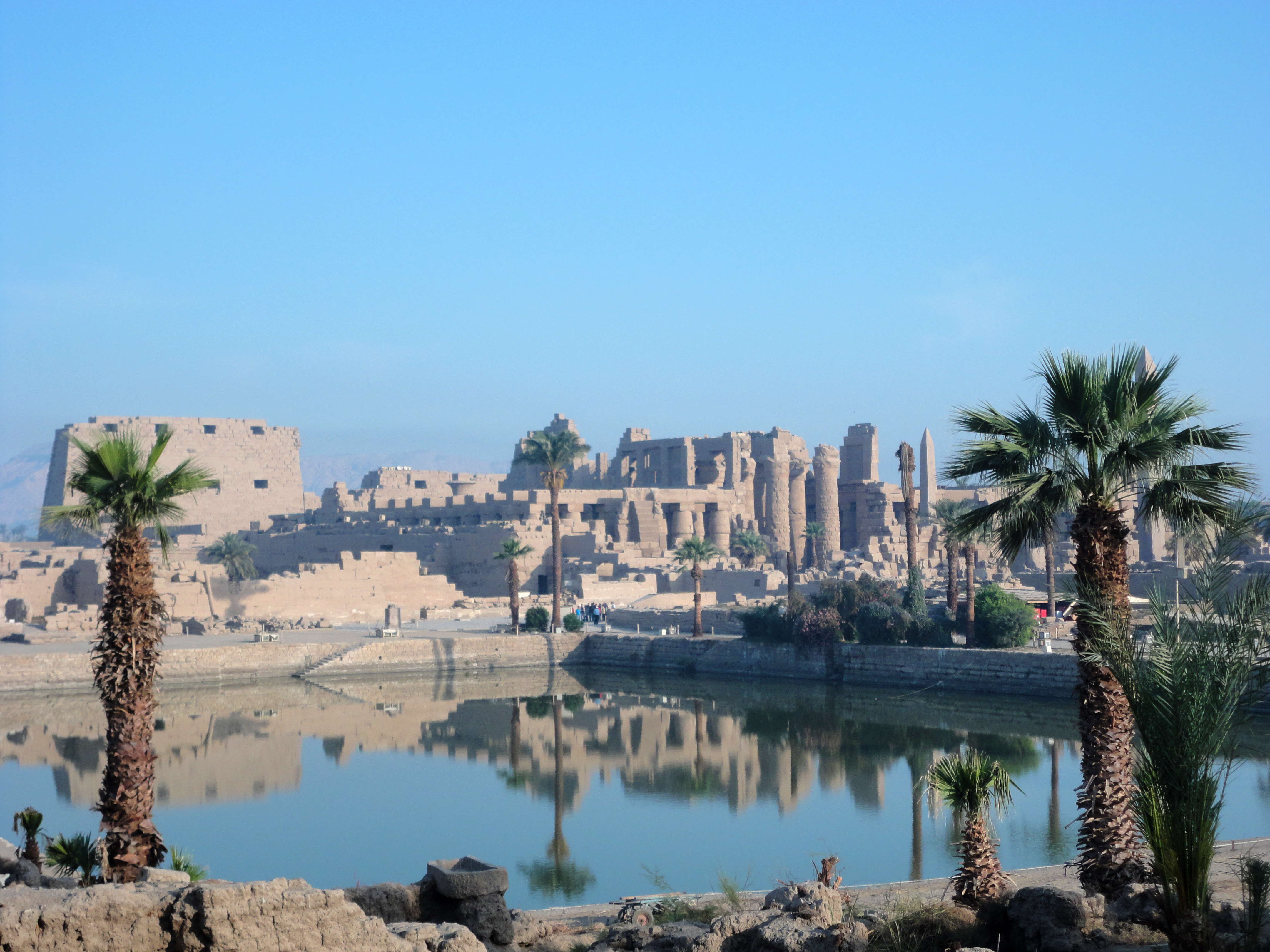 Ancient ruins of the Karnak Temple complex in Luxor, Egypt, viewed across a large reflecting pool with palm trees in the foreground. The stone columns, pylons, and obelisks are reflected in the still water under a clear blue sky.
