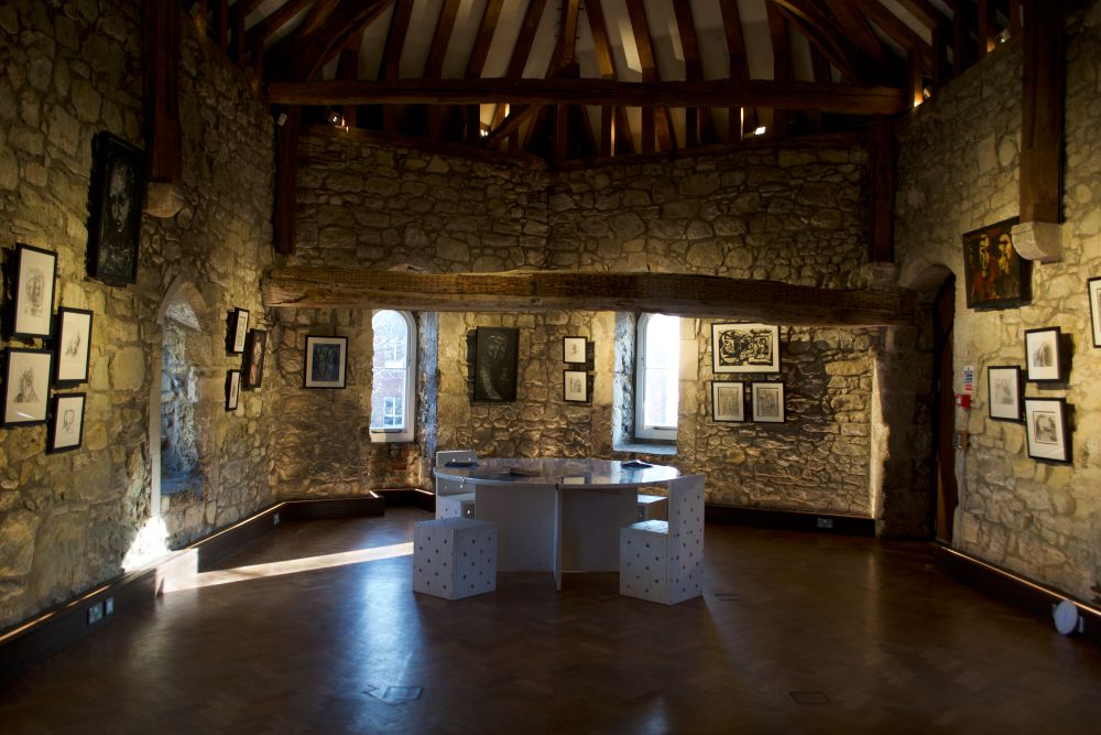 An art gallery room with stone walls and a high timbered ceiling displays framed drawings and prints along the walls. In the center, a circular table with stools sits on a polished wooden floor, illuminated by warm lighting.