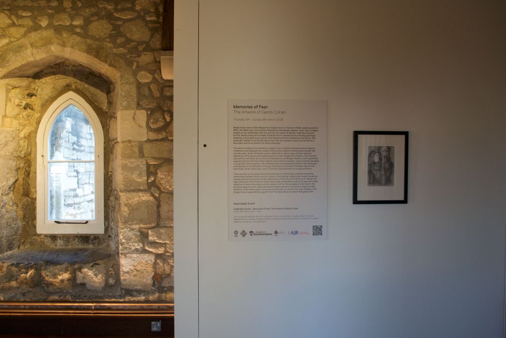 A stone‑walled gallery space displays a framed artwork and a wall panel of exhibition text beside an arched window. Sunlight comes through the window, highlighting the textured stone surface.