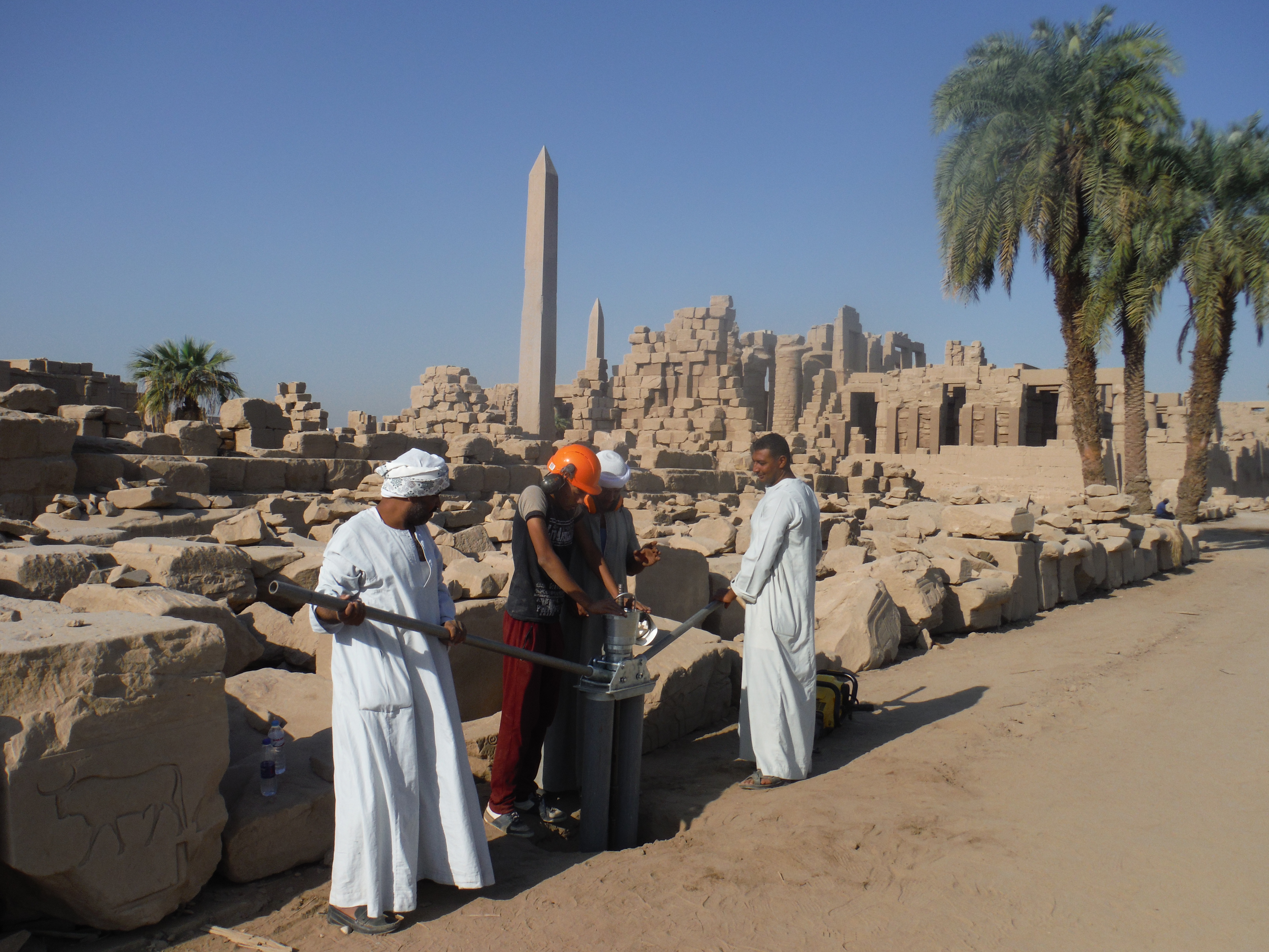 Four men work with equipment among the ruins of the Karnak Temple complex in Luxor, Egypt. Two of them wear traditional white garments, while another wears a hard hat. Ancient stone blocks, palm trees, and tall obelisks rise in the background under a clear blue sky.