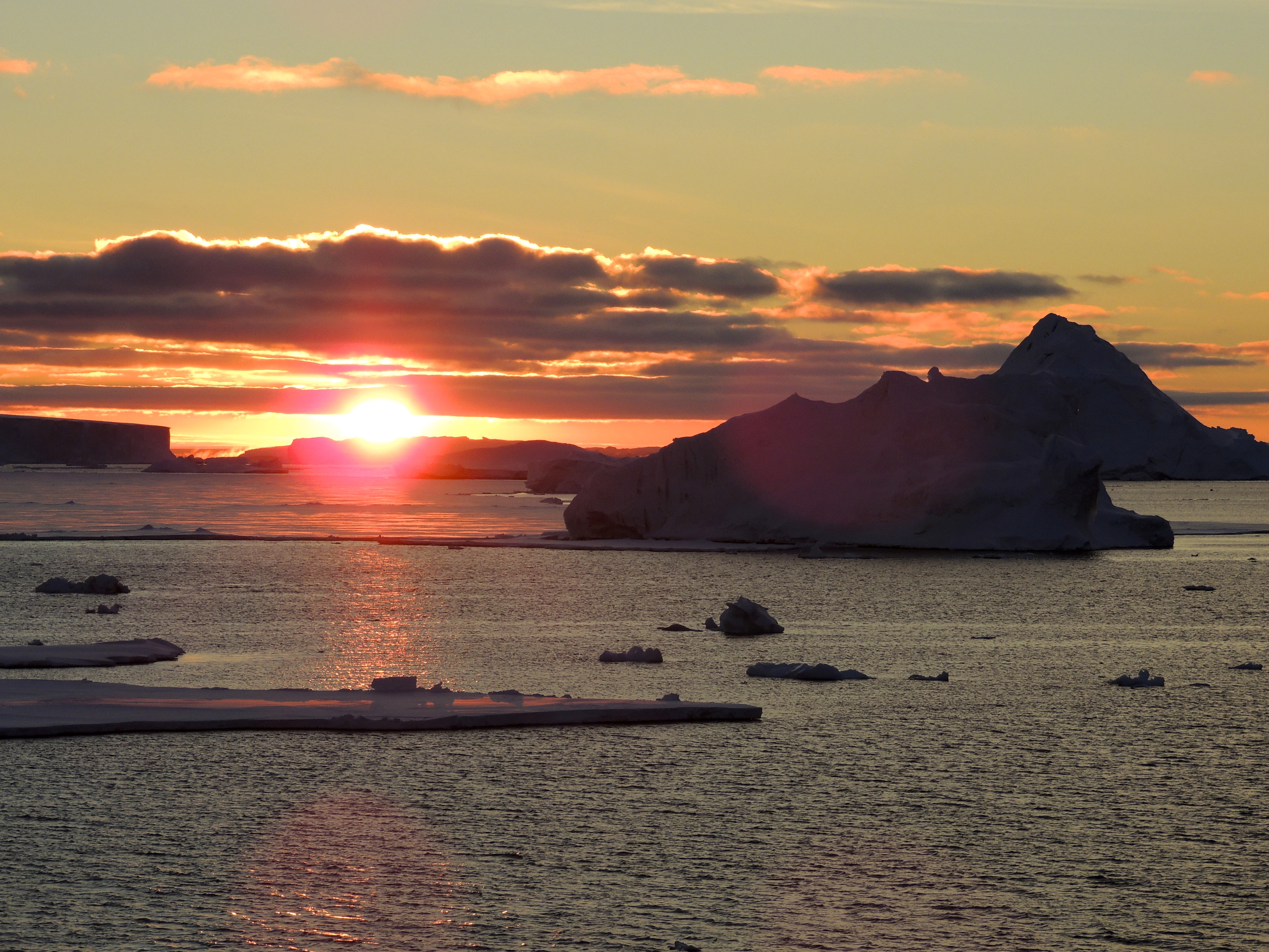 A dramatic polar sunset over icy waters, with the sun dipping below the horizon and casting a warm orange and pink glow across the sky and sea. Large icebergs and ice floes float in the dark water, and a massive, jagged iceberg dominates the right side of the image. Low clouds stretch across the sky, reflecting the fading sunlight.