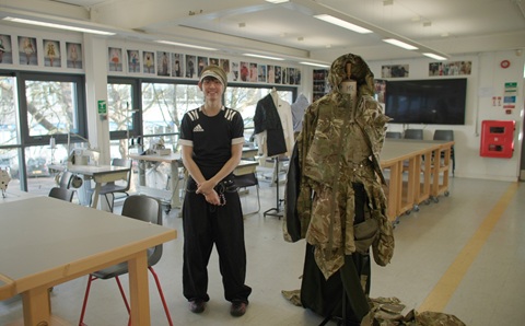 Young man standing inside a large room with desks and sewing machines. He is standing next to a full size mannequin that has camouflage clothing on it.