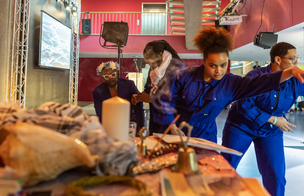 Performers in blue jumpsuits gather around a table filled with symbolic objects such as candles, fabric, and natural materials, while a projection screen and stage setup appear in the background. The scene captures a moment of movement and interaction within a brightly lit performance space.