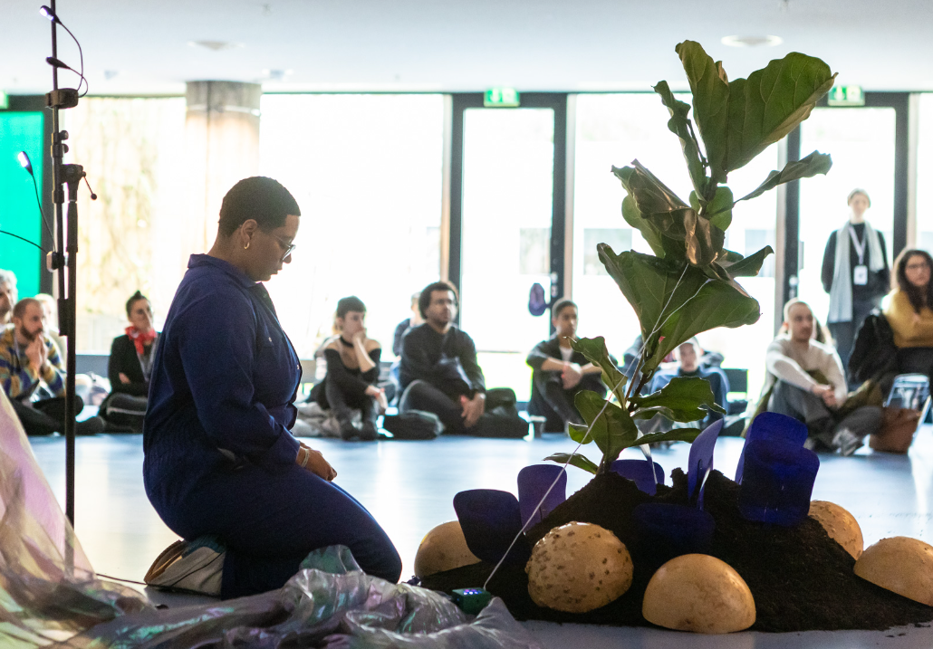 A performer in a blue jumpsuit kneels beside an arrangement of soil, stones, plants, and blue glass vessels, while an audience seated on the floor and chairs observes from behind. Natural light fills the room through large windows.