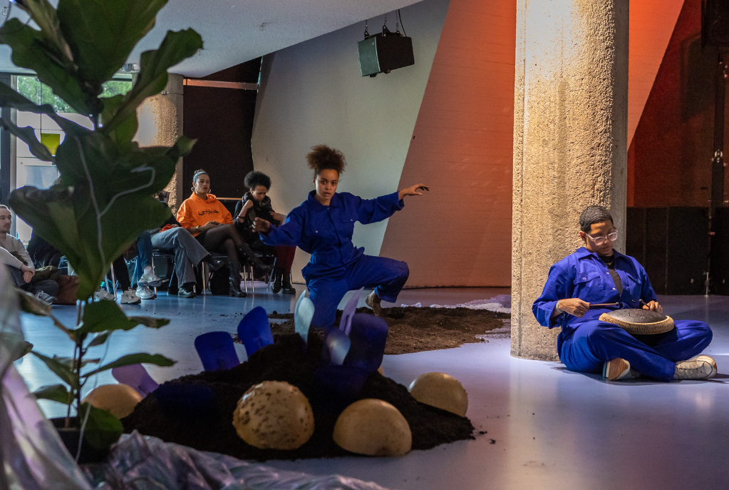 Two performers in blue jumpsuits move around a sculptural installation of soil, stones, and blue glass pieces, while others sit and watch from the surrounding space. The setting is a spacious indoor area with concrete pillars and soft, atmospheric lighting.