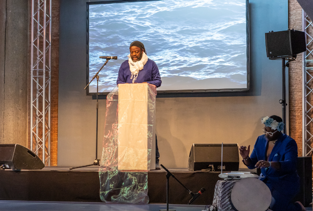 A performer stands behind a podium draped in translucent fabric, speaking into a microphone in front of a large projection of ocean waves. Another performer sits on the stage nearby holding a drum-like object.