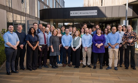 A group of about 20 adults standing together facing the camera. They are stood outside on a paved area, in front of a building