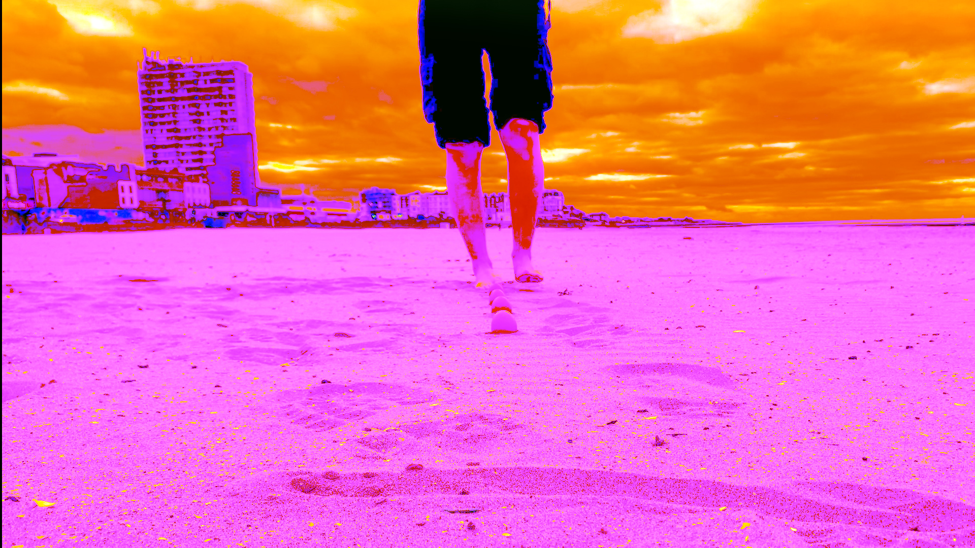 A vibrant, color-enhanced photo shows a person's legs walking barefoot on a beach, leaving footprints in the sand, with three eggs placed in a line in front of them. The sky is an intense orange, and the surrounding buildings are slightly distorted in bright pink and purple tones.