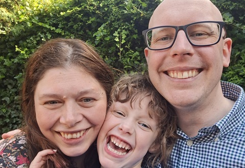 Woman with long brown hair, young girl, and a man in glasses, all smiling. They are in front of a green hedge.