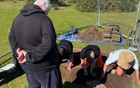 Three people on their knees digging in an area of soil, with a man in a black jacket standing watching